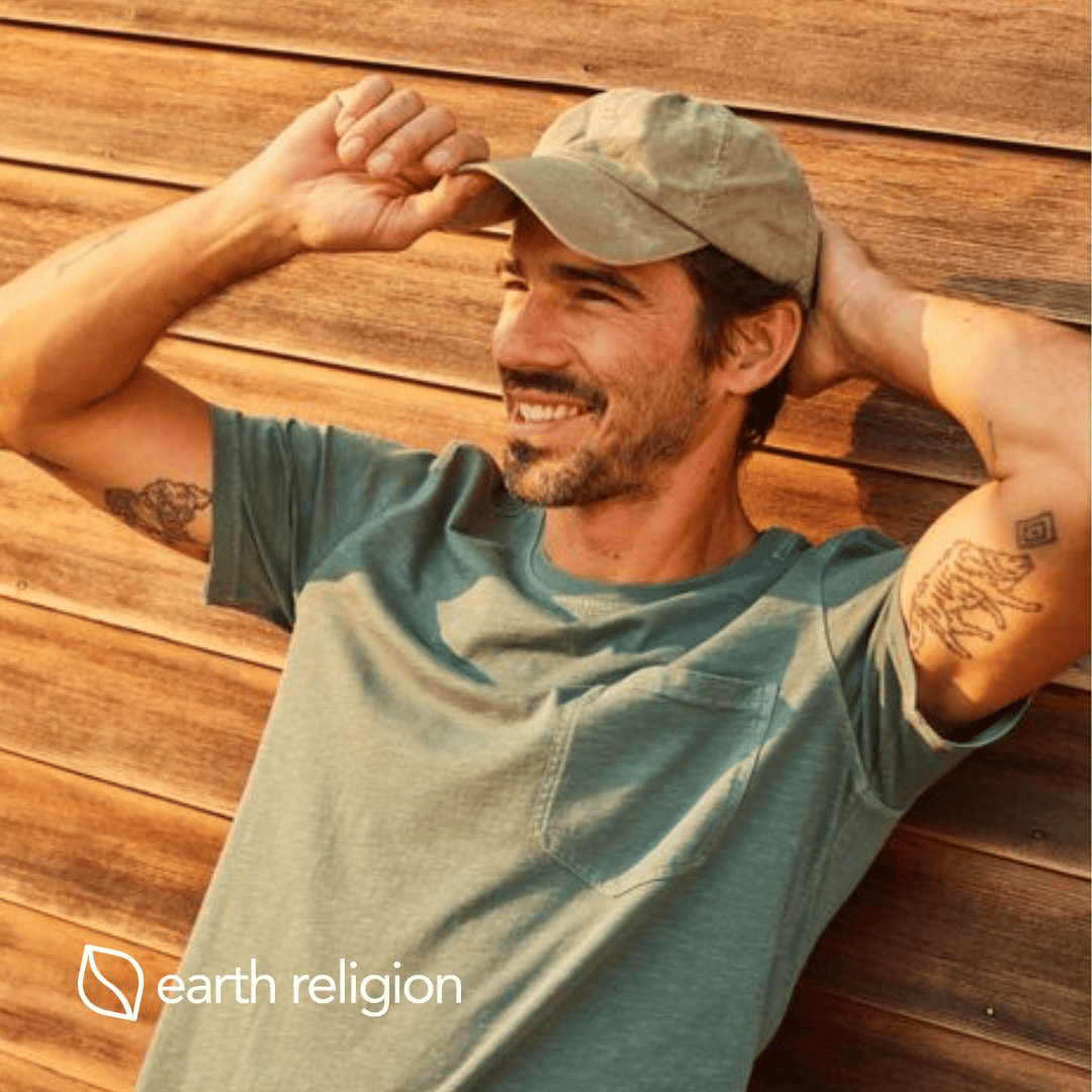 A man smiling, resting against a wall with his hands on his hat as if he just put it on.